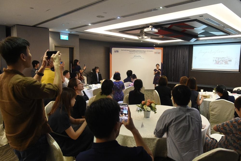 Jia Ying sharing her story at the ceremony as audience listen intently and take videos.