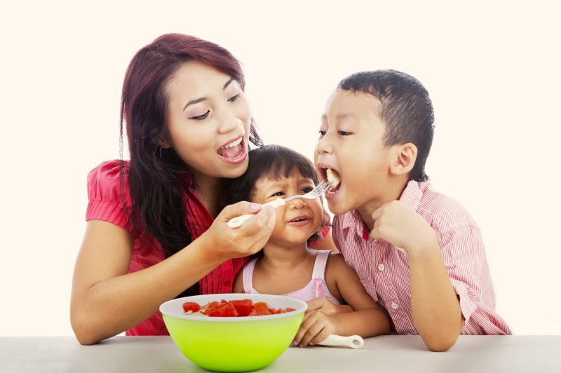 Experiencing Language - A Multisensory Approach to Language Learning 3 A mother feeding her boy a piece of fruit as a girl between them looks on