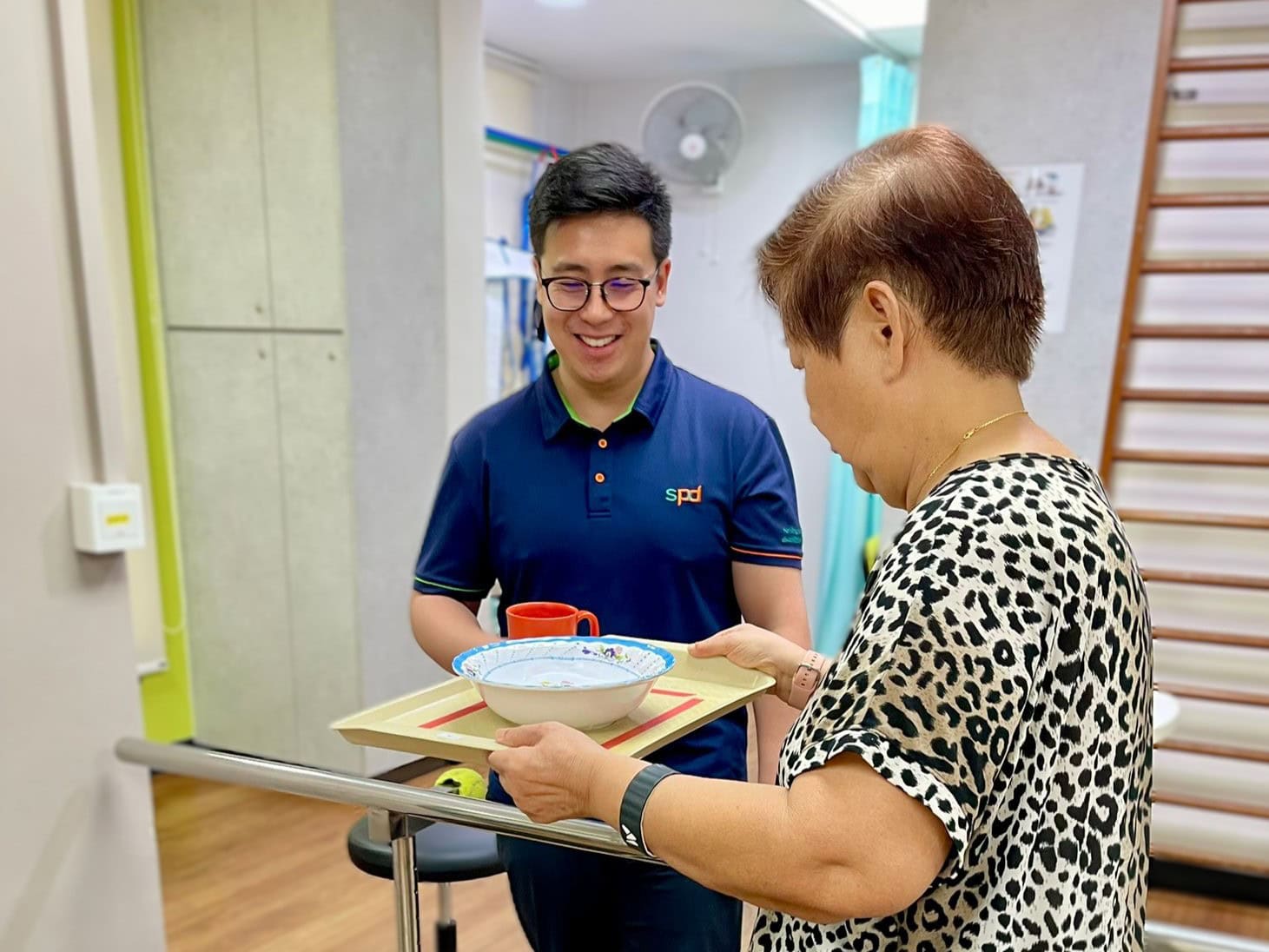 Power of Care – Growing with Purpose 1 Joel looks on as a senior practises walking with a tray of crockery