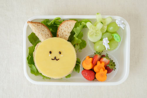 A lunch box with fruit, vegetables and open face sandwich with a rabbit face