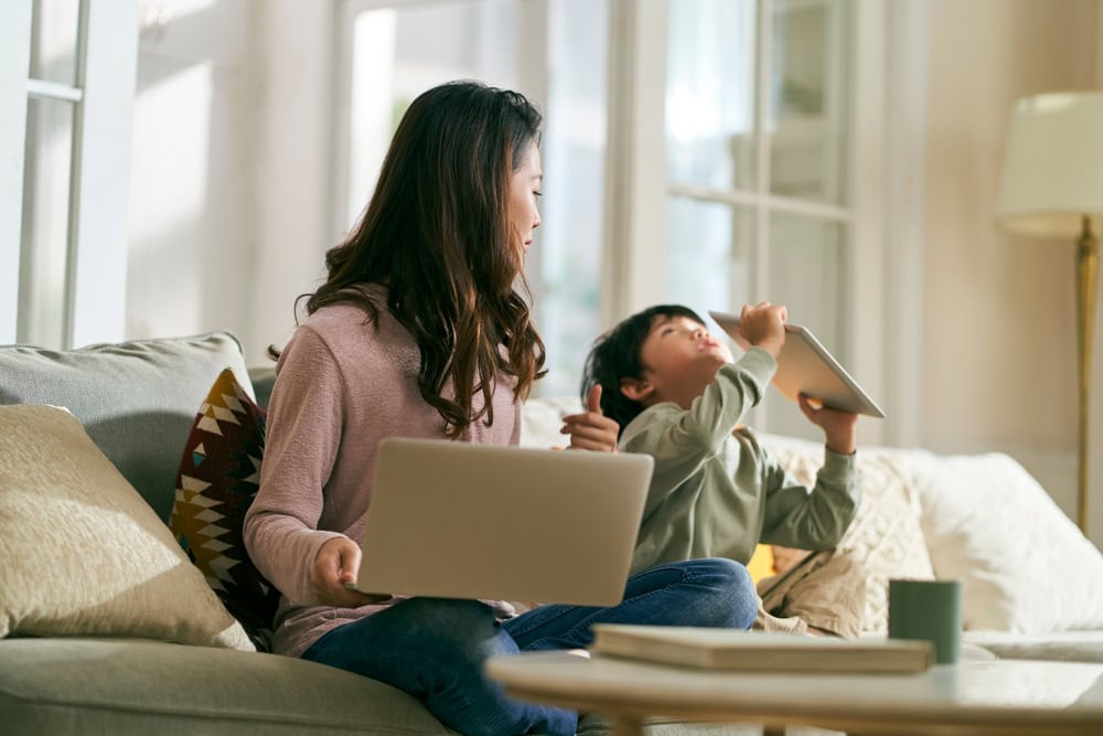 A mother sitting on the sofa working on her laptop, with her is her child who is distracted from his tablet