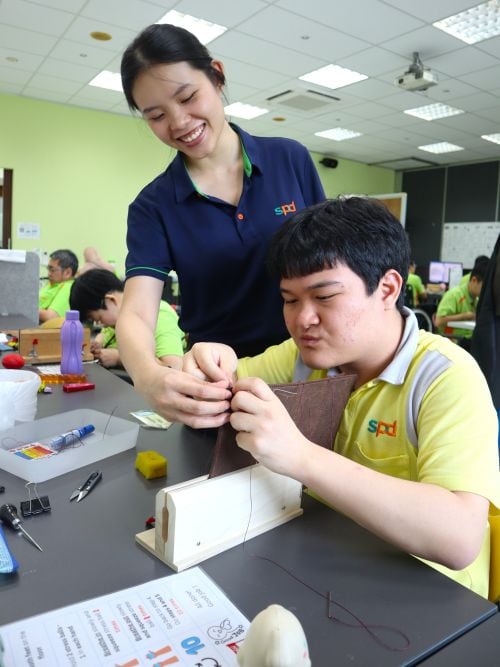 An S P D workshop supervisor guiding Joel in sewing leather products