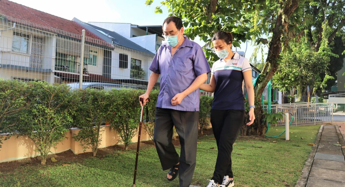 S P D professional supporting an elderly man walking with a walking stick by holding onto his elbow