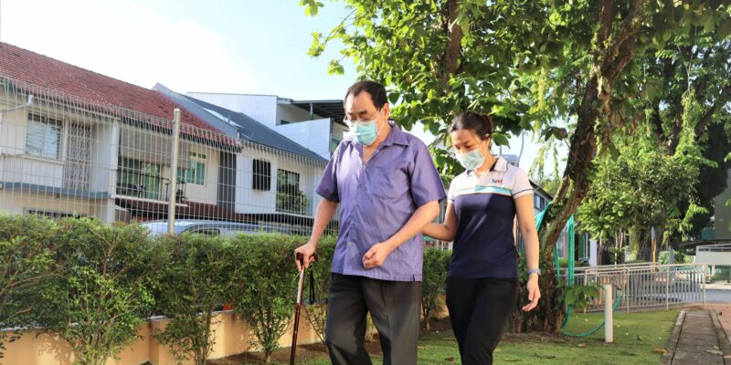 S P D professional supporting an elderly man walking with a walking stick by holding onto his elbow