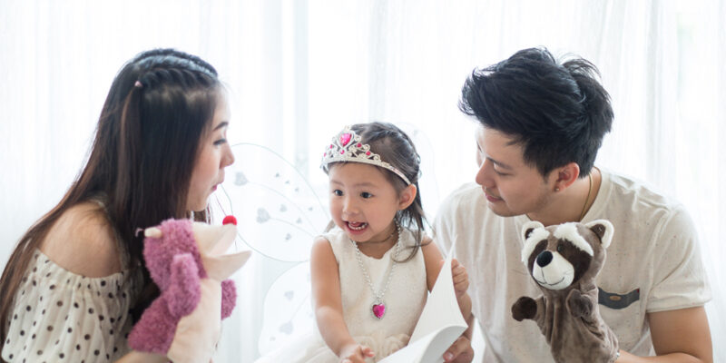 Mother and father holding up puppets with a girl pointing at book