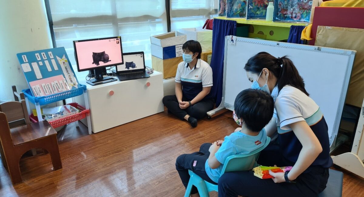 A boy seated looking at a monitor and laptop screen with 2 SPD staff seated beside