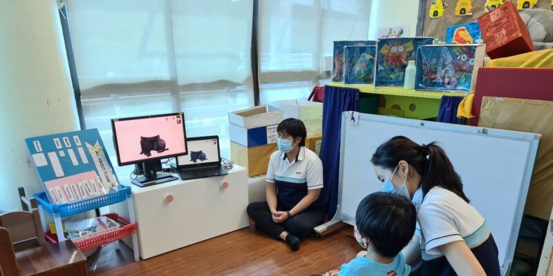 A boy seated looking at a monitor and laptop screen with 2 SPD staff seated beside