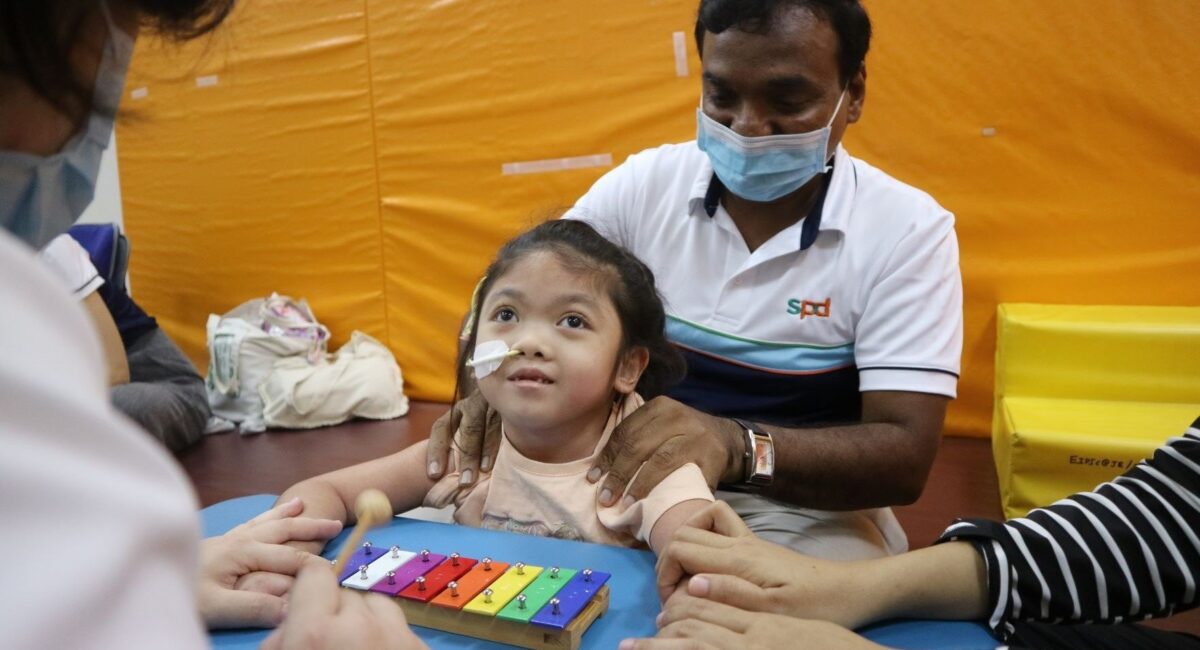 A smiling Syaza facing a therapist playing on the xylophone whilst physiotherapist Chandra helps her with sitting upright