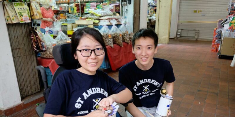 A lady on wheelchair and a man on her right side, holding up tin cans for fundraising while facing the camera with a smile.