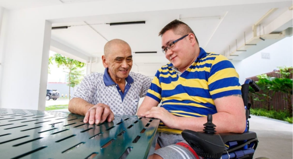 Mr Kua sitting on a bench at the void deck with his stepson Mr Lim, who is using a wheelchair
