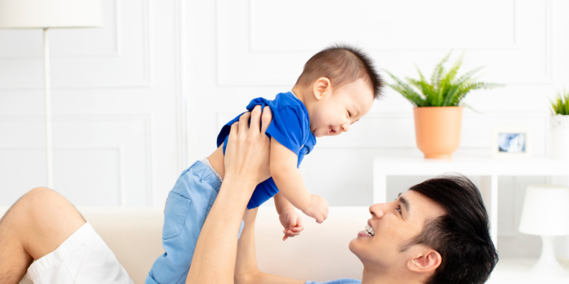 A dad lying on the sofa while lifting his young son up in the air