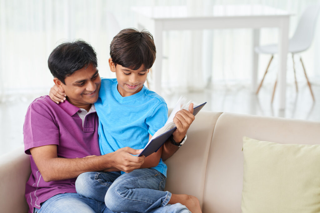 A young boy sitting on his father's lap as they read a book on the sofa