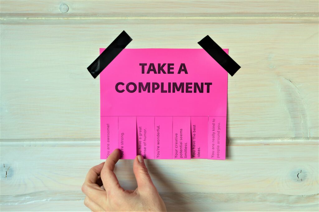 Pink stickers with text of compliments pasted on a wooden wall. The text in image reads “Take a compliment".