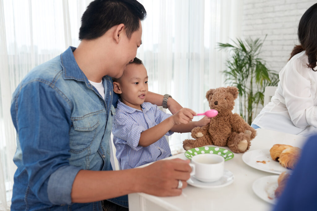 A father putting his arm around his son's shoulder as he watches his son feed a toy bear in a game of pretend play
