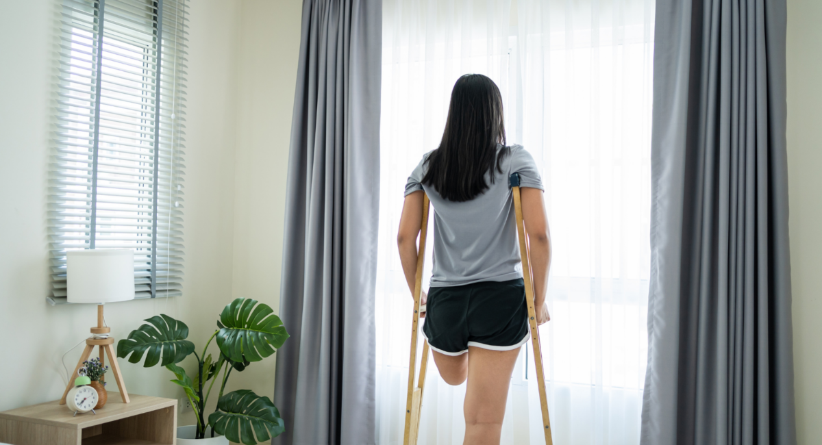Young female amputee standing supported with crutches while looking out of the bedroom window