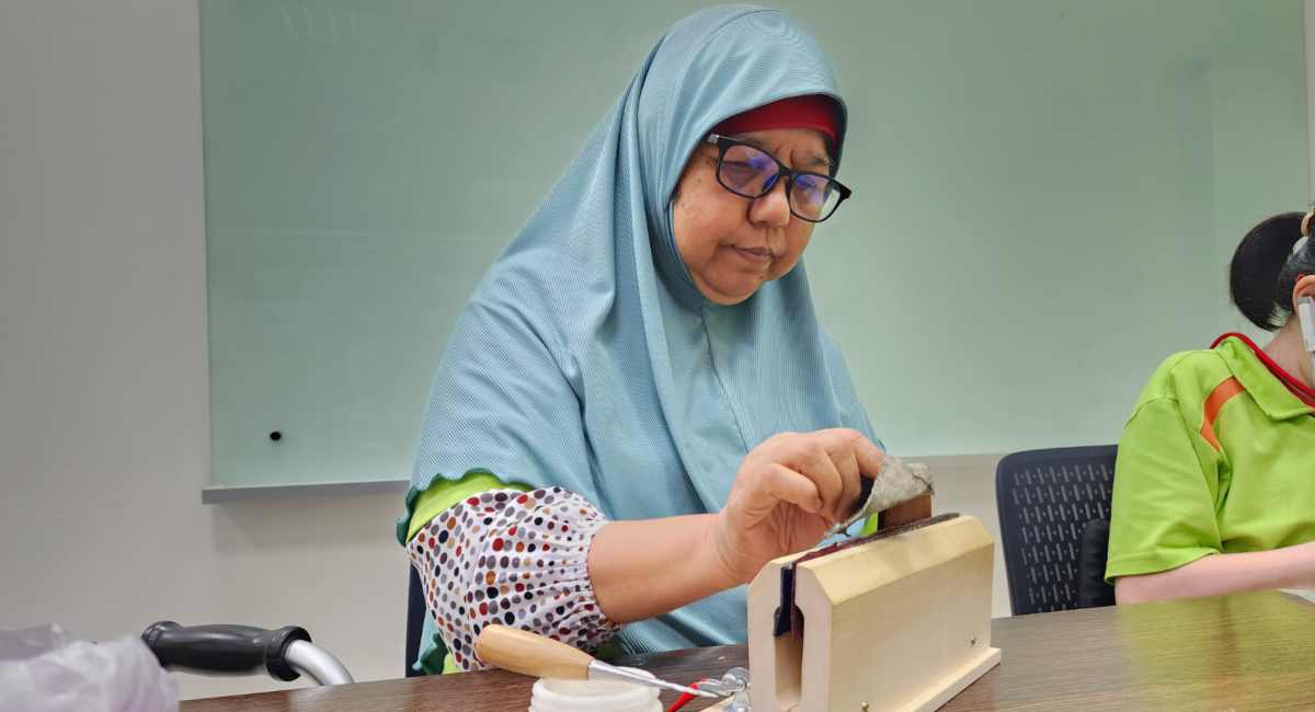 Mdm Hamidah sewing a leather piece that is held steady by a wooden clamp