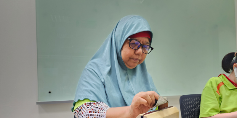 Mdm Hamidah sewing a leather piece that is held steady by a wooden clamp