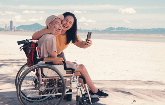 A lady taking a selfie with a male teenager who is using a wheelchair at a beach. Both smiling.
