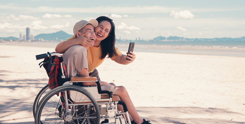 A lady taking a selfie with a male teenager who is using a wheelchair at a beach. Both smiling.