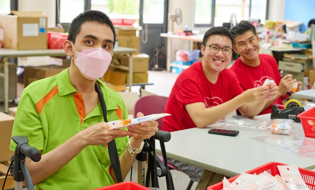 2 Red Hat volunteers packing products at the S P D Sheltered Workshop with trainee Alvin