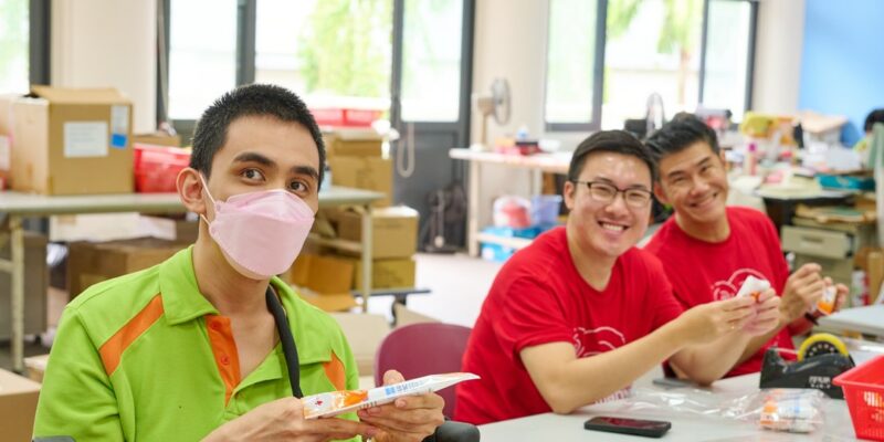 2 Red Hat volunteers packing products at the S P D Sheltered Workshop with trainee Alvin