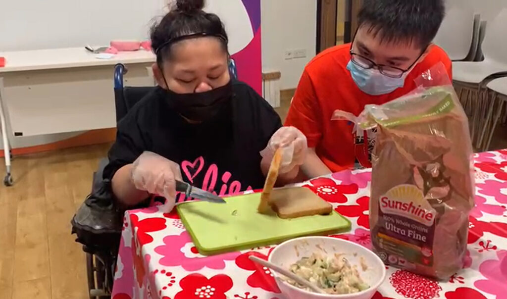 A wheelchair user preparing a meal with sliced bread and salad. A young man in spectacles looks on.