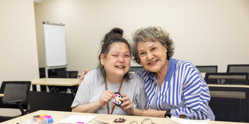 Veronica and mum Mdm Lydia smiling, a table of craft items scattered on the table they are at