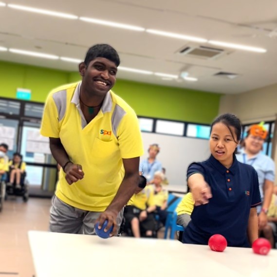 Puvan waiting excitedly to roll a blue ball across the table. An SPD training officer points at a general direction for him to throw at whilst a volunteer and his peers look on.