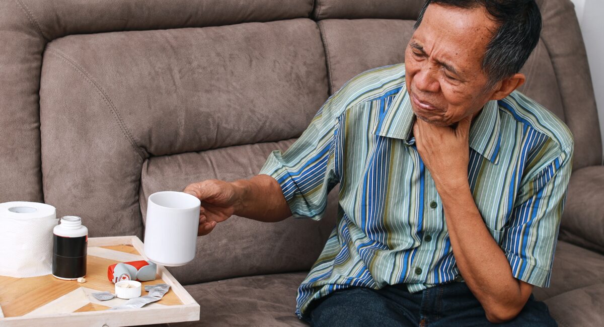 An elderly man holding his throat and looking pained whilst holding up a mug