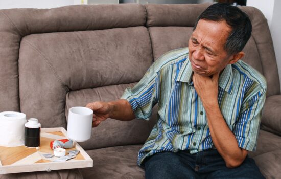 An elderly man holding his throat and looking pained whilst holding up a mug
