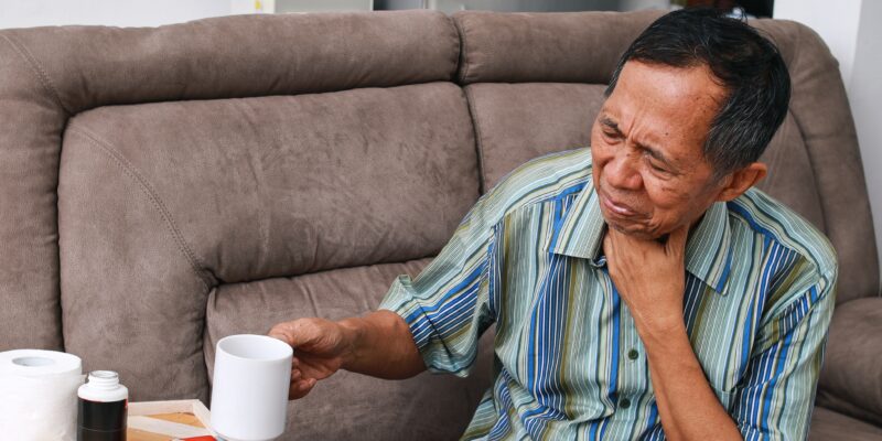 An elderly man holding his throat and looking pained whilst holding up a mug