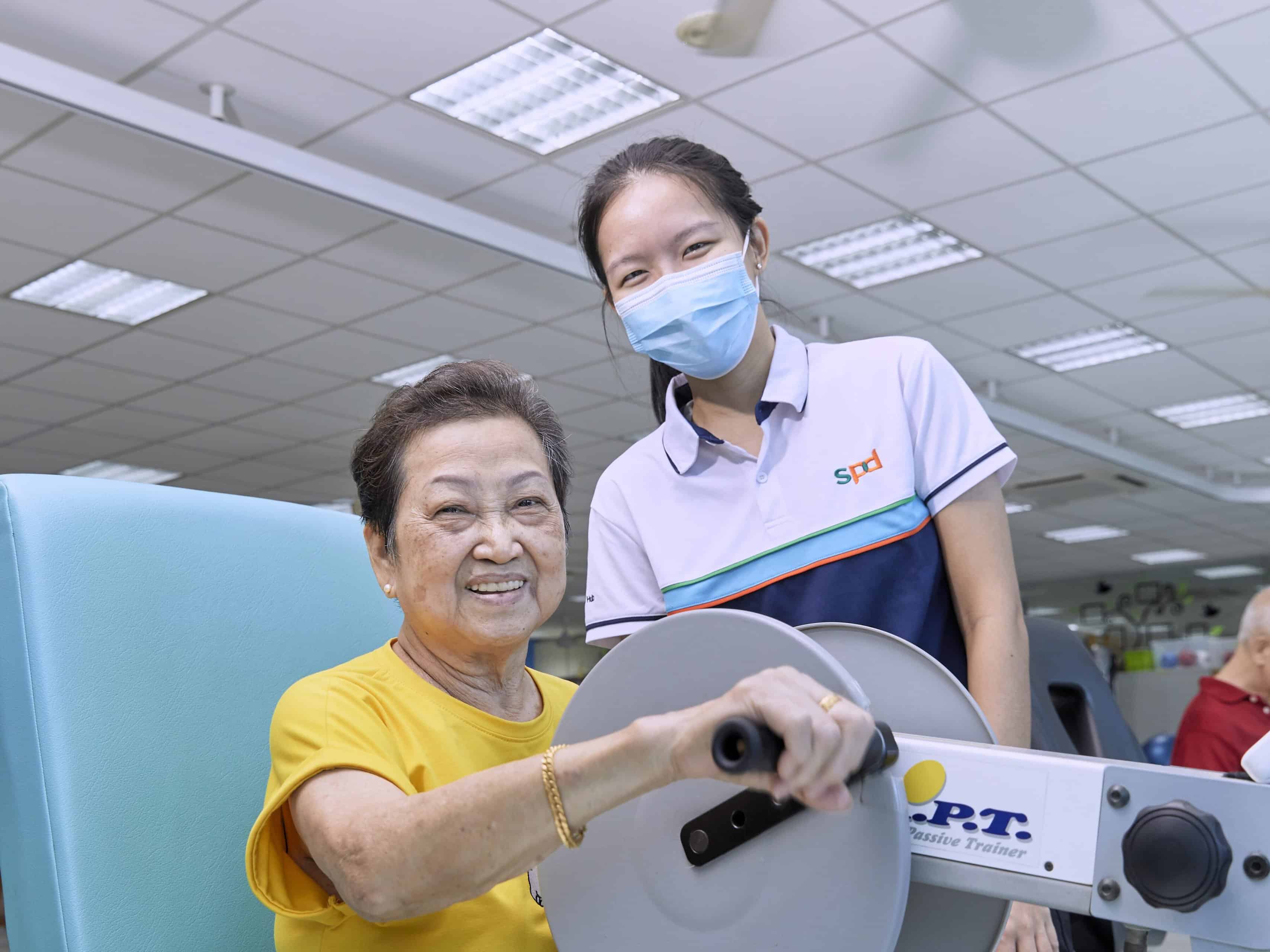 A senior working on a handcycle machine as an S P D therapist looks on