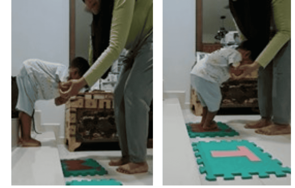 A caregiver supporting a boy by his elbow to learn to jump from the last steps of the stairs to a colourful mat on the floor.