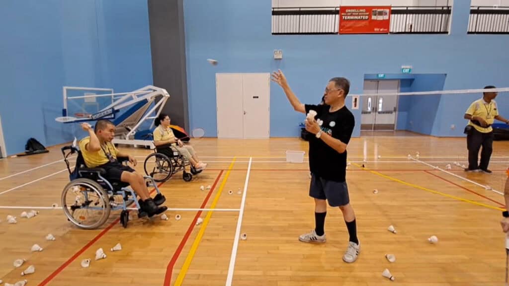 A volunteer throws a shuttlecock towards D W W client Ernest, a wheelchair user, who looks to hit it with his badminton racket