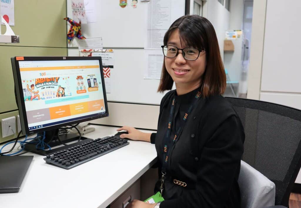 Partnerships manager Adeline in black long sleeve blouse, sitting in front of a monitor showing S P D Ability Walk and Run webpage