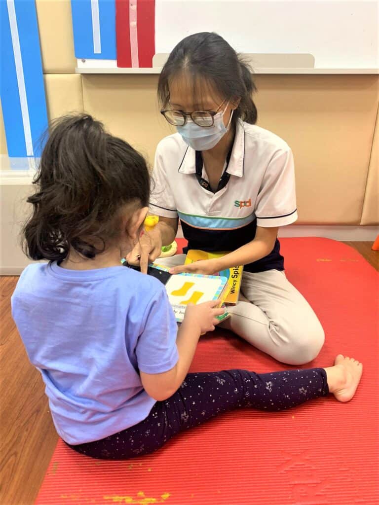 Teacher Mei Yi sitting on a red mat in a classroom setting, working on an activity with a young girl