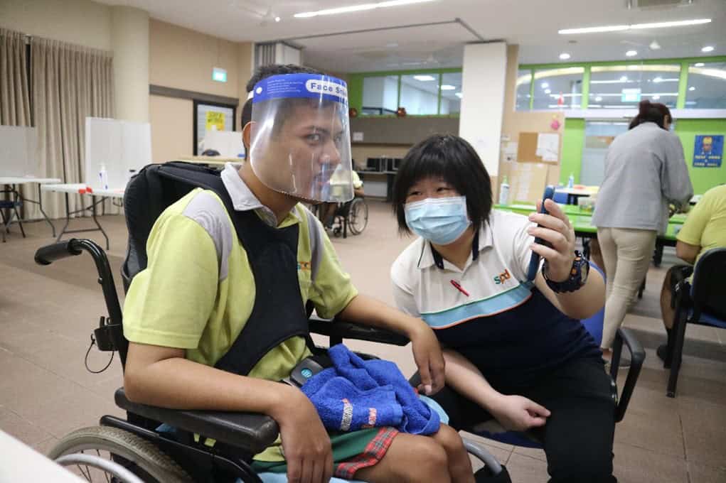 Rena holding up and showing her handphone to a day activity centre client. He is in a wheelchair and is wearing a face shield.