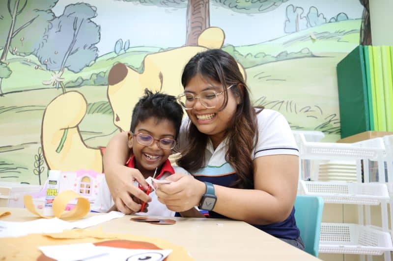 Teacher Isma guiding a boy in cutting paper, both are smiling broadly.