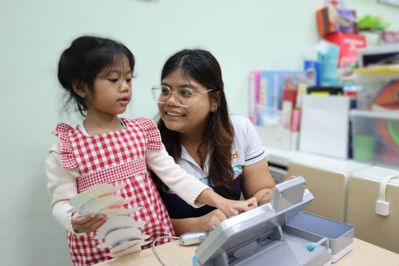 A young girl is typing on a toy register whilst Teacher Isma looks at her tenderly.