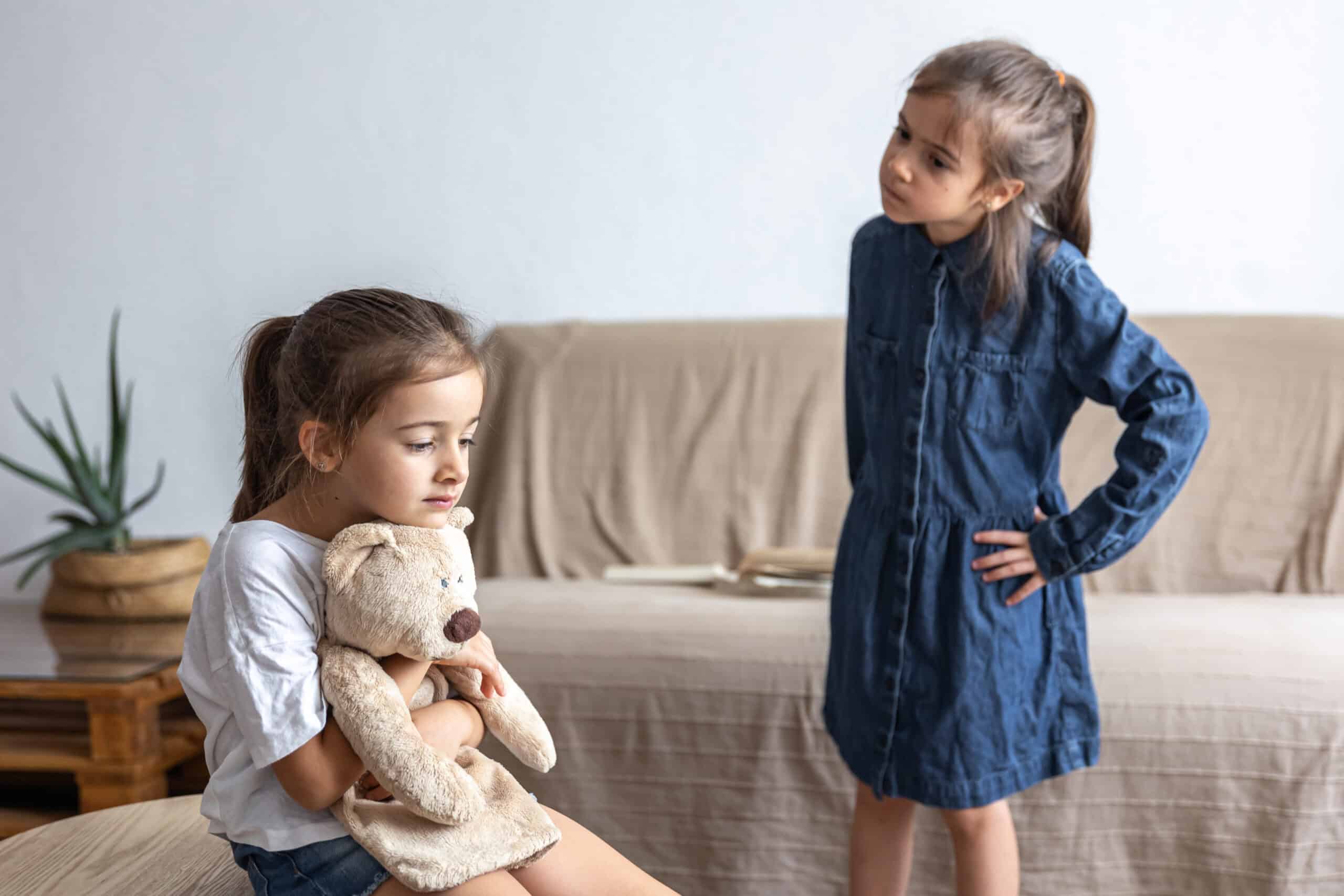 A girl in white sitting down and hugging a soft toy whilst another girl in blue with hands a the waist looks on in annoyance