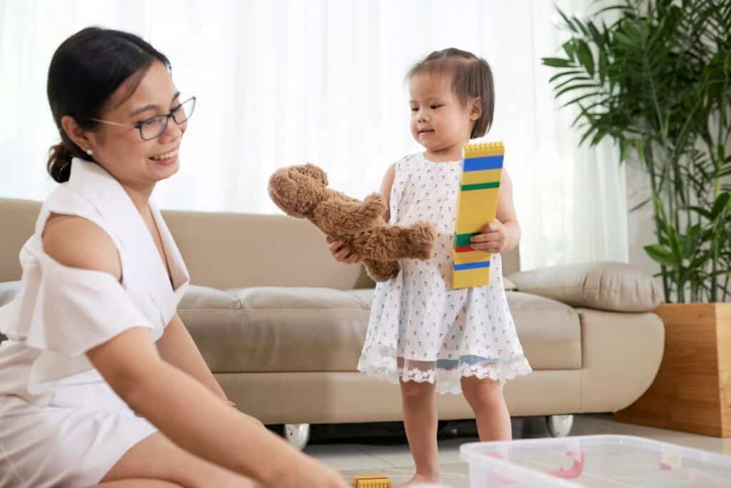 A mother with her young daughter who is holding on to a toy bear and blocks