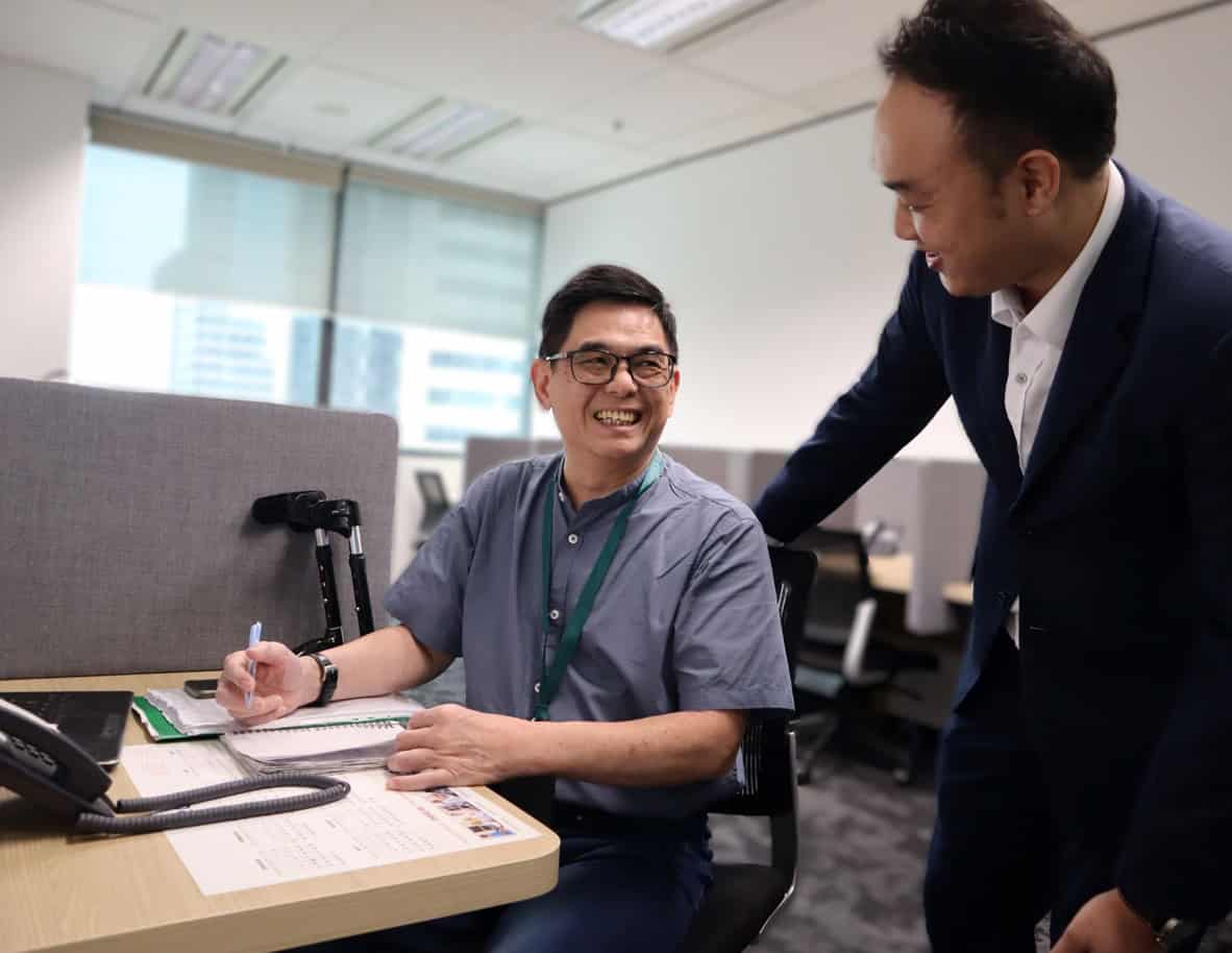 A man in a suit chatting with a person seated at his desk and his crutches placed against a cubicle divider