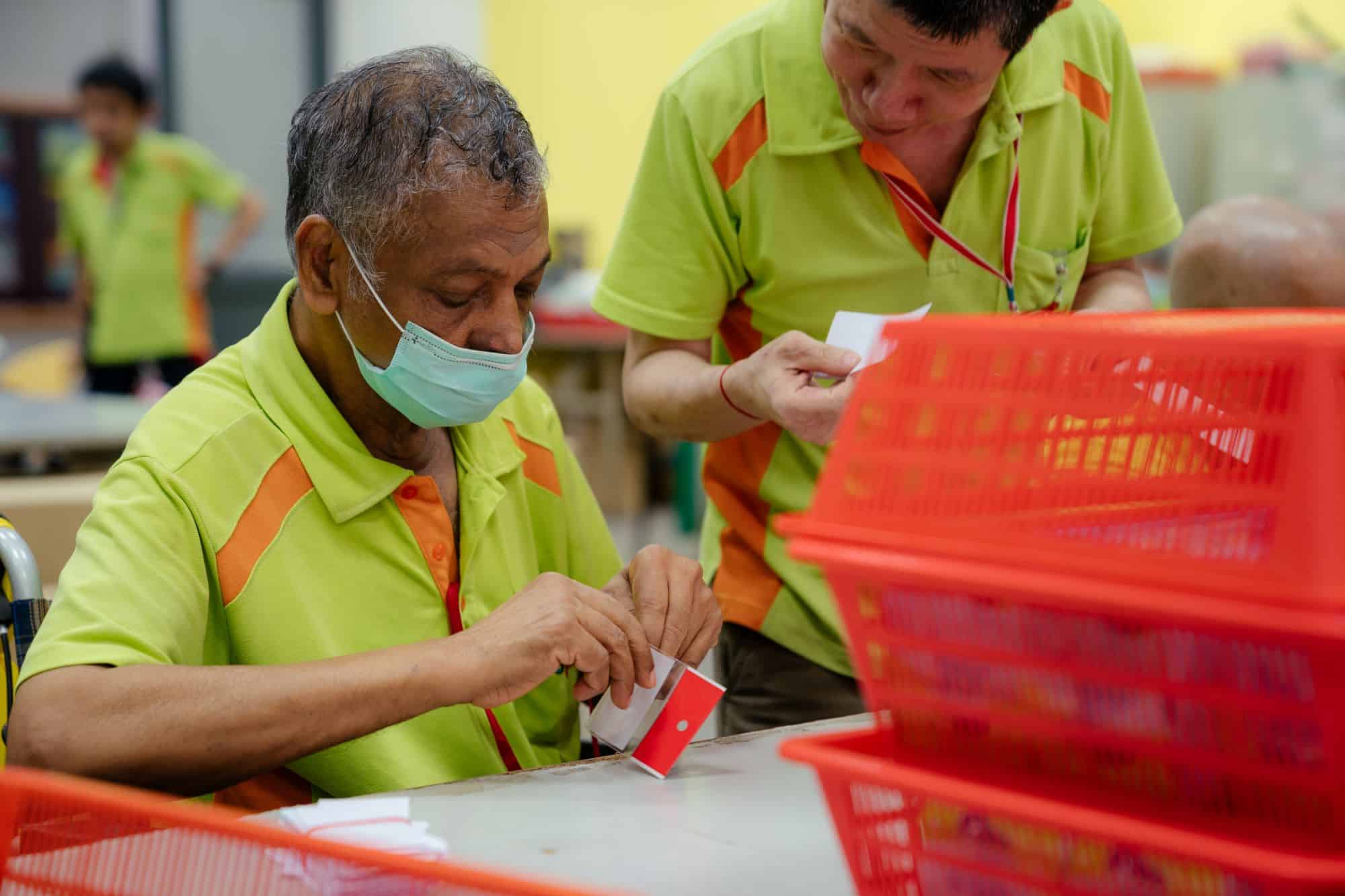 A Sheltered Workshop trainee working on working on the adhesive of a plastic package as another trainee looks on.