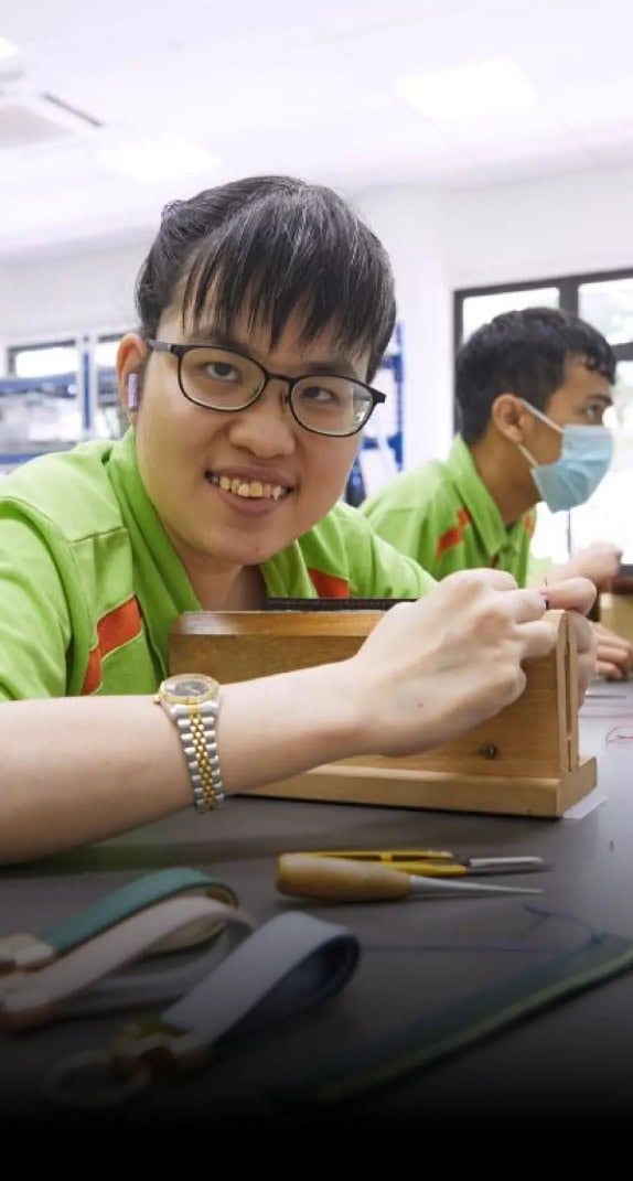 A young female S P D trainee sewing some leather products and smiling.