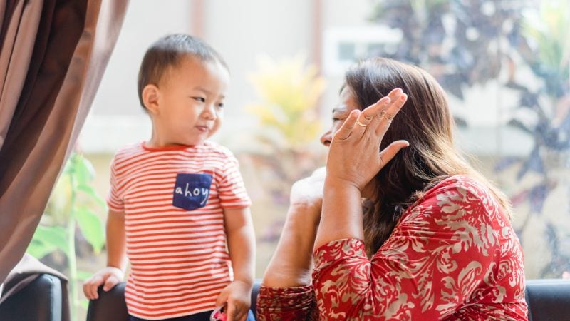 A mother playing peekaboo with a boy, both at eye level to each other