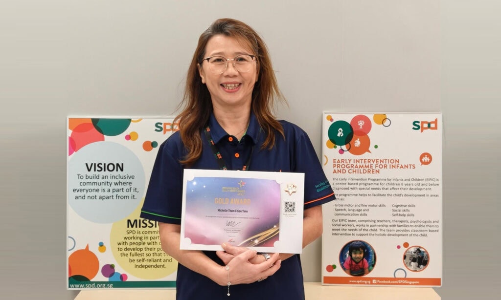 Michelle holding her award, smiling and in front of S P D's mission and early intervention programme description board.