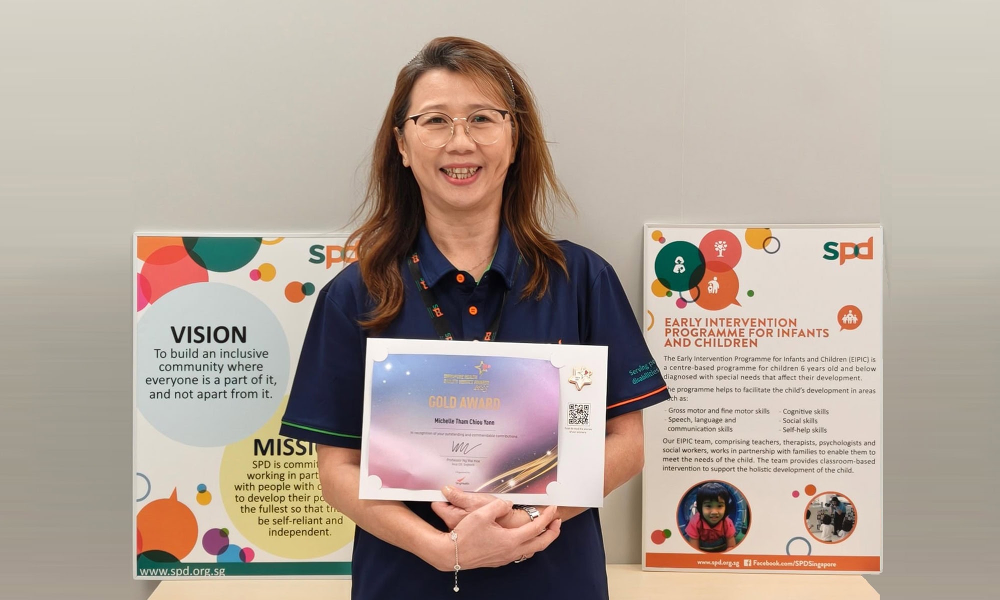 Michelle holding her award, smiling and in front of S P D's mission and early intervention programme description board.
