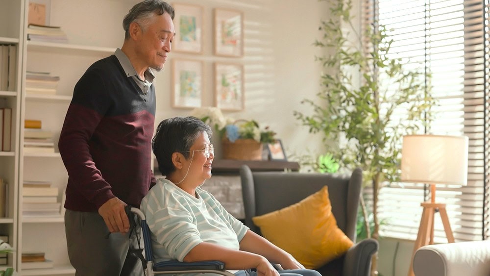 An elderly man holding onto the handles of a wheelchair which an elderly woman is seated in, both smiling.