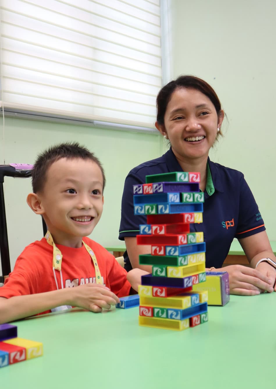 Home 5 A boy smiling widely with his teacher in a classroom, both look to be in a middle of a game of jenga.