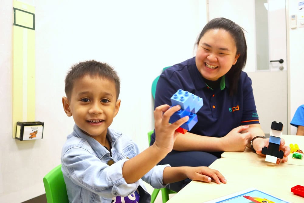 A young boy playing with some multi-coloured plastic bricks as his teacher looks on
